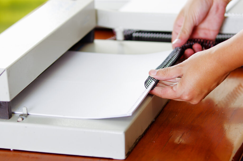 Hands inserting a plastic coil binding into a stack of printed pages using a binding machine.